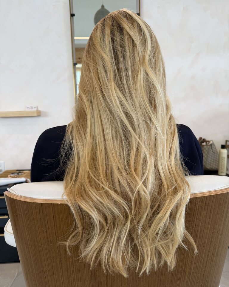 French balayage result — long warm-blonde hair, photographed against a pink-plaster wall in the salon