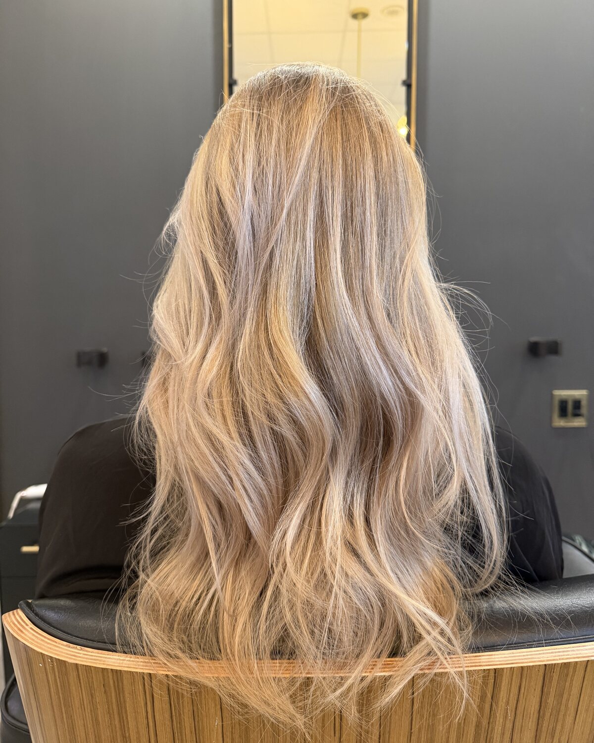 Long, soft ash-blonde waves, photographed from behind in the salon against a lit mirror